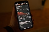 A hand holding a smartphone displaying a stock market app with a graph showing a downward trend in the Dow Jones Industrial Average. The app interface shows detailed stock information including numerical values and recent stock news updates.