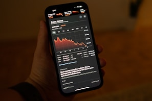 A hand holding a smartphone displaying a stock market app with a graph showing a downward trend in the Dow Jones Industrial Average. The app interface shows detailed stock information including numerical values and recent stock news updates.