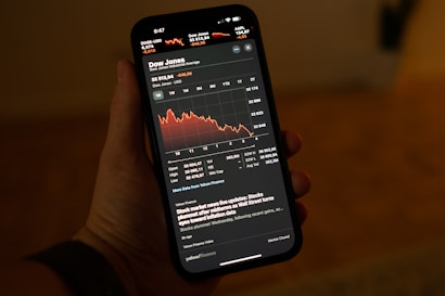 A hand holding a smartphone displaying a stock market app with a graph showing a downward trend in the Dow Jones Industrial Average. The app interface shows detailed stock information including numerical values and recent stock news updates.