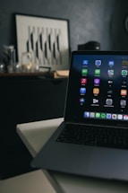 A laptop with various application icons displayed on the screen is positioned on a table in a dimly lit room. The background features a framed artwork with black and white geometric patterns resting on a dark piece of furniture, alongside several glass jars.
