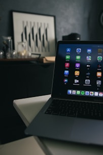 A laptop with various application icons displayed on the screen is positioned on a table in a dimly lit room. The background features a framed artwork with black and white geometric patterns resting on a dark piece of furniture, alongside several glass jars.
