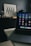 A laptop with various application icons displayed on the screen is positioned on a table in a dimly lit room. The background features a framed artwork with black and white geometric patterns resting on a dark piece of furniture, alongside several glass jars.