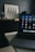 A laptop with various application icons displayed on the screen is positioned on a table in a dimly lit room. The background features a framed artwork with black and white geometric patterns resting on a dark piece of furniture, alongside several glass jars.