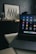 A laptop with various application icons displayed on the screen is positioned on a table in a dimly lit room. The background features a framed artwork with black and white geometric patterns resting on a dark piece of furniture, alongside several glass jars.
