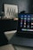 A laptop with various application icons displayed on the screen is positioned on a table in a dimly lit room. The background features a framed artwork with black and white geometric patterns resting on a dark piece of furniture, alongside several glass jars.