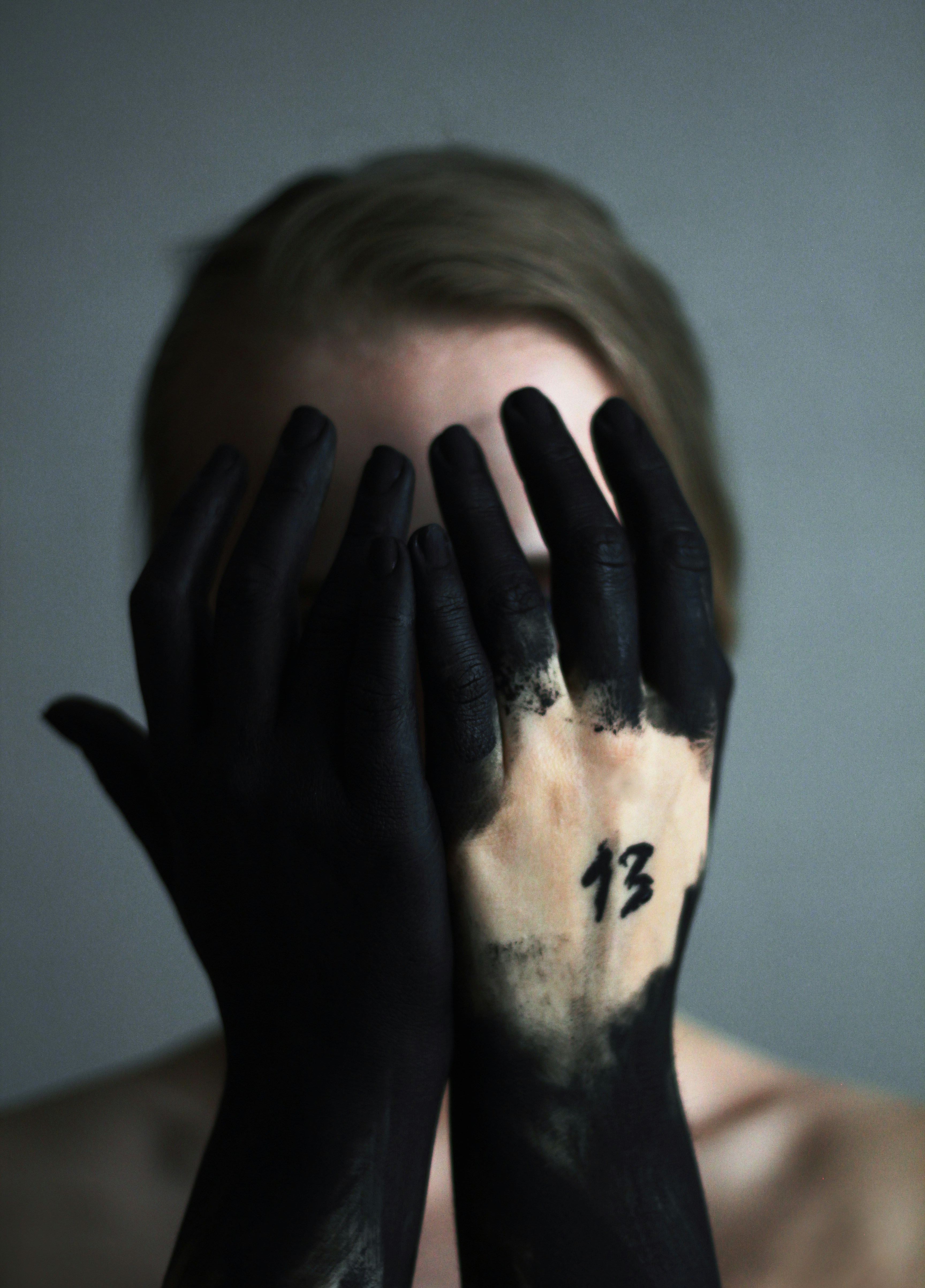 A young woman covers her face with her hands. Her hands are covered in black paint. The number 13 on her hand. Thirteen