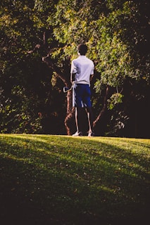 A person stands on a grassy hill, holding a golf club, with tall green trees in the background. The lighting creates a contrast between the dark shadows and the illuminated areas.