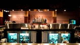 A well-stocked bar with various liquor bottles displayed prominently and a selection of beer taps in the center. Behind the counter, there are glass-door refrigerators filled with bottled drinks. The bar has a polished metal surface, and the surrounding decor features patterned wallpaper and warm lighting, creating a cozy atmosphere.