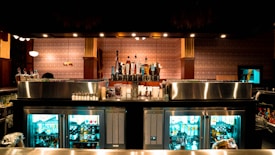A well-stocked bar with various liquor bottles displayed prominently and a selection of beer taps in the center. Behind the counter, there are glass-door refrigerators filled with bottled drinks. The bar has a polished metal surface, and the surrounding decor features patterned wallpaper and warm lighting, creating a cozy atmosphere.
