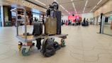 A luggage trolley is placed in an airport terminal with several people in the background. The trolley is stacked with a variety of suitcases and has a sculptural element of a cat. The terminal features modern design elements, such as wooden paneling and overhead lights. People are queuing at a counter in the distance.