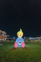A large, colorful inflatable sculpture of a cartoon-like character is situated on a grassy field at night. The character has a yellow head with pink cheeks and a single blue strand of hair, a blue outfit with white patterns, and bright pink feet. The area is dimly lit by surrounding buildings and streetlights, creating a whimsical nighttime atmosphere.