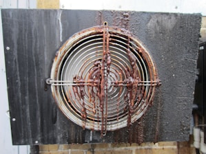 A metal vent or fan cover with circular grille covered in a thick layer of rust and grime. The grille is mounted on a black wall and the wall has visible stains. The surface appears wet and corroded.