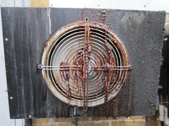 A metal vent or fan cover with circular grille covered in a thick layer of rust and grime. The grille is mounted on a black wall and the wall has visible stains. The surface appears wet and corroded.