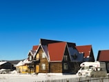 A wooden house with red roofs is situated in a snowy landscape. The structure has multiple triangular roof sections and is accompanied by two white vehicles parked nearby. A picket fence outlines the property, and the sky is clear and blue.