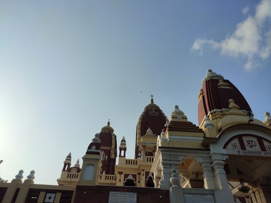 A Hindu temple with intricate architecture featuring domes and spires, adorned with religious symbols and carvings. The structure is primarily in red and cream colors, set against a clear blue sky with a few clouds.