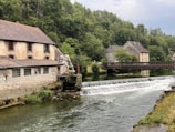 A tranquil riverside scene featuring a stone building with a tiled roof next to a flowing river. A small dam creates a gentle waterfall in the water. Across the river, a wooden footbridge connects to another stone building surrounded by lush greenery. Trees line the background, creating a serene and peaceful countryside atmosphere.