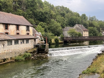 A tranquil riverside scene featuring a stone building with a tiled roof next to a flowing river. A small dam creates a gentle waterfall in the water. Across the river, a wooden footbridge connects to another stone building surrounded by lush greenery. Trees line the background, creating a serene and peaceful countryside atmosphere.