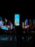 A vibrant urban night scene featuring numerous digital billboards with brightly lit advertisements. The central billboard showcases a nature-themed image with underwater elements like corals and fish. Surrounding billboards display a variety of images and text. Below, small illuminated water fountains create colorful light reflections on the ground.