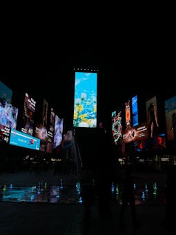 A vibrant urban night scene featuring numerous digital billboards with brightly lit advertisements. The central billboard showcases a nature-themed image with underwater elements like corals and fish. Surrounding billboards display a variety of images and text. Below, small illuminated water fountains create colorful light reflections on the ground.