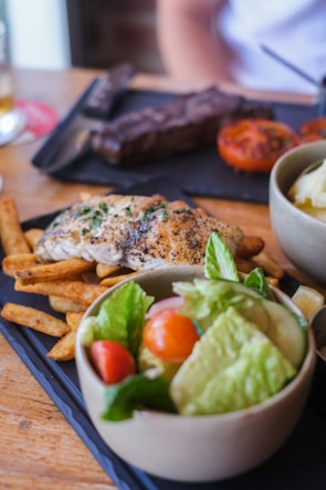 A meal consisting of grilled chicken breast seasoned with herbs, served with crispy fries. A bowl of fresh salad with lettuce, cherry tomatoes, and cucumber accompanies the dish. In the background, there is a steak and grilled tomato on a black slate serving tray.