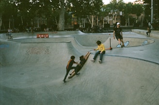 A skatepark scene with several children playing. Two children are at the bottom of a concrete ramp while another appears to be helping one up. A child in a yellow shirt is seated at the top of a ramp with a scooter. Trees and a building are visible in the background.
