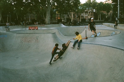 A skatepark scene with several children playing. Two children are at the bottom of a concrete ramp while another appears to be helping one up. A child in a yellow shirt is seated at the top of a ramp with a scooter. Trees and a building are visible in the background.