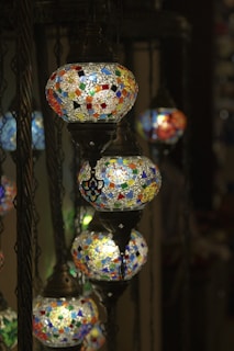 A series of intricately designed mosaic lamps hang from dark metal chains. Each lamp is adorned with colorful, geometric patterns made from small, stained glass pieces, casting a warm and inviting glow.