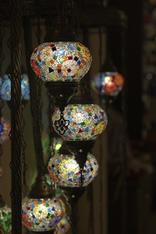 A series of intricately designed mosaic lamps hang from dark metal chains. Each lamp is adorned with colorful, geometric patterns made from small, stained glass pieces, casting a warm and inviting glow.