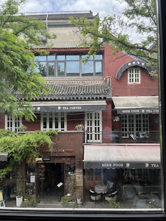 A charming two-story building with oriental architectural elements appears nestled amid lush green foliage. The facade is painted a deep red with traditional roofing. Large windows on the upper floor are complemented by awnings advertising tea, coffee, and beer food. White-framed doors and windows add a classic touch. Outdoor seating is arranged on the lower level, hinting at a cozy cafe or restaurant ambiance.