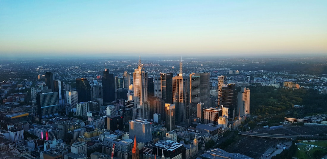 Aerial view of a city skyline during sunset, with modern skyscrapers and high-rise buildings casting long shadows. The cityscape is surrounded by a mix of green spaces and urban infrastructure, extending into the horizon. The sky features a gradient from soft blue to warm golden hues.