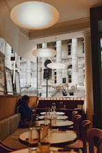 A cozy dining area within a restaurant featuring elegant round tables set with glass bottles and glasses. Warm lighting from overhead lamps creates a welcoming atmosphere. A person is seated on a cushioned bench, looking at their phone, and large windows offer a view of a historic building facade outside.