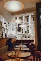 A cozy dining area within a restaurant featuring elegant round tables set with glass bottles and glasses. Warm lighting from overhead lamps creates a welcoming atmosphere. A person is seated on a cushioned bench, looking at their phone, and large windows offer a view of a historic building facade outside.