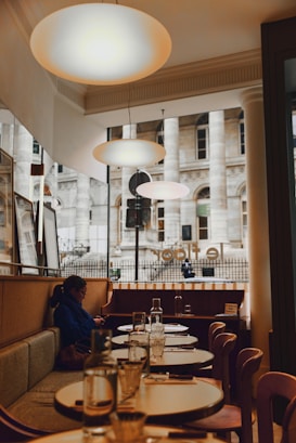 A cozy dining area within a restaurant featuring elegant round tables set with glass bottles and glasses. Warm lighting from overhead lamps creates a welcoming atmosphere. A person is seated on a cushioned bench, looking at their phone, and large windows offer a view of a historic building facade outside.