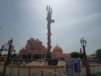 An ornate temple with intricate carvings and a large trident structure in the foreground. The temple features multiple domes and decorative elements. The sky is clear and there are some signs and lampposts around the structure.
