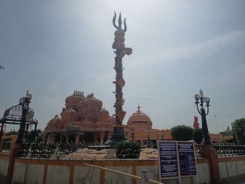 An ornate temple with intricate carvings and a large trident structure in the foreground. The temple features multiple domes and decorative elements. The sky is clear and there are some signs and lampposts around the structure.