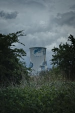 A large industrial cooling tower is surrounded by dense vegetation and trees. The sky above is overcast with dark clouds, giving a moody atmosphere. The cooling tower has a weathered appearance with patches of discoloration.