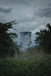 A large industrial cooling tower is surrounded by dense vegetation and trees. The sky above is overcast with dark clouds, giving a moody atmosphere. The cooling tower has a weathered appearance with patches of discoloration.