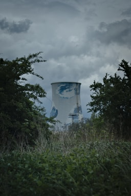A large industrial cooling tower is surrounded by dense vegetation and trees. The sky above is overcast with dark clouds, giving a moody atmosphere. The cooling tower has a weathered appearance with patches of discoloration.