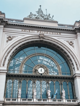 An elaborate architectural structure with a large arched window and intricate details. A clock is centrally located below the arch, and statues are placed along the ledge beneath the window. The building facade has detailed ornamentation and the words 'KELETI PALYAUDVAR' inscribed above the arch. Three flags are also visible.