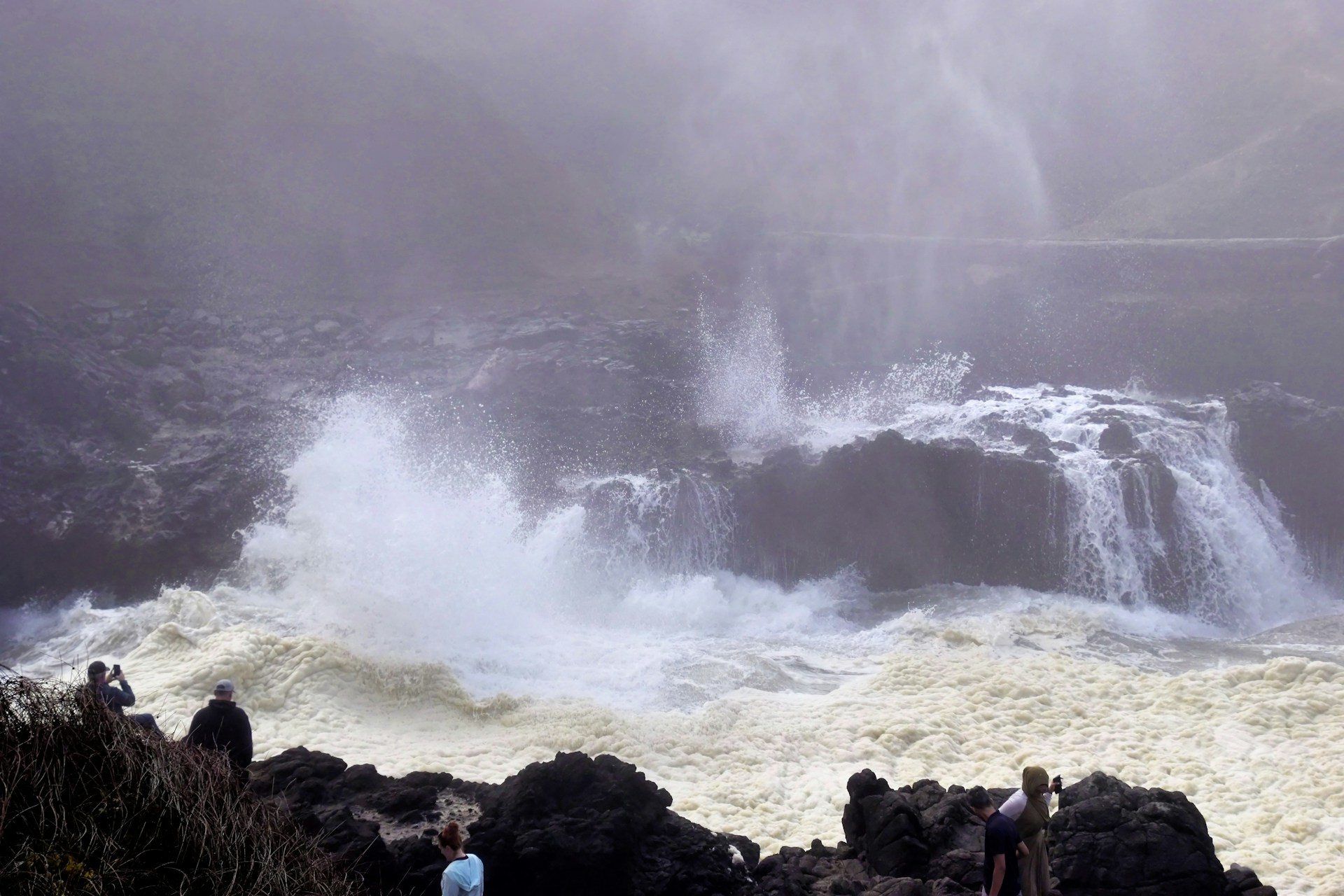 A wide shot of a group of photographers gathered on a rocky coastline, focused on the crashing waves under a dramatic sky.