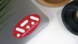 A laptop with a red sticker featuring white geometric shapes and text on its surface. To the right, there is a camera lens with detailed markings. A wooden bowl or holder is partially visible beside the lens. On the left, green plant leaves add a natural touch.