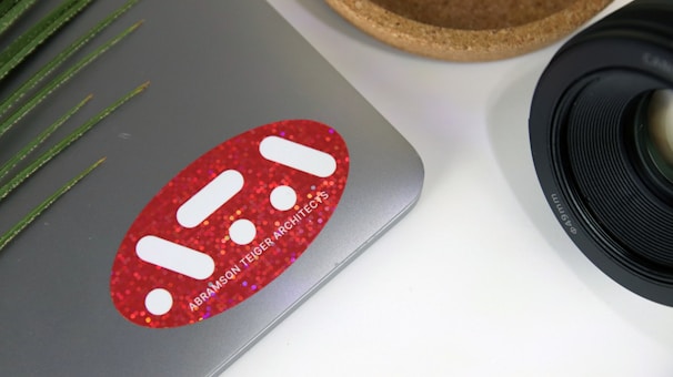 A laptop with a red sticker featuring white geometric shapes and text on its surface. To the right, there is a camera lens with detailed markings. A wooden bowl or holder is partially visible beside the lens. On the left, green plant leaves add a natural touch.