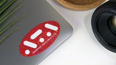 A laptop with a red sticker featuring white geometric shapes and text on its surface. To the right, there is a camera lens with detailed markings. A wooden bowl or holder is partially visible beside the lens. On the left, green plant leaves add a natural touch.