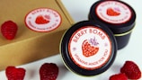 Several tins labeled 'Berry Bomb Vitamins Made for You' are displayed alongside fresh raspberries. The labels feature a stylized image of a strawberry. A cardboard box is also partially visible.