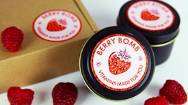 Several tins labeled 'Berry Bomb Vitamins Made for You' are displayed alongside fresh raspberries. The labels feature a stylized image of a strawberry. A cardboard box is also partially visible.