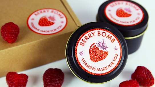 Several tins labeled 'Berry Bomb Vitamins Made for You' are displayed alongside fresh raspberries. The labels feature a stylized image of a strawberry. A cardboard box is also partially visible.