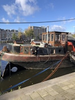 A weathered boat is docked by the side of a canal with a person working on it. The boat features a rustic cabin with windows and various tools and equipment scattered around. The background shows a row of traditional multi-story buildings with trees and a tall modern building further away.
