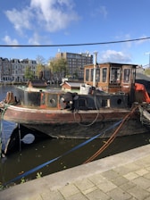 A weathered boat is docked by the side of a canal with a person working on it. The boat features a rustic cabin with windows and various tools and equipment scattered around. The background shows a row of traditional multi-story buildings with trees and a tall modern building further away.