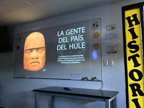 A classroom setting with a projection of an Olmec head statue displayed on a white screen. The text on the screen reads 'LA GENTE DEL PAÍS DEL HULE' along with a description below, mentioning the significance of the Olmec culture as the mother culture. On the right side, there is a yellow sign with the word 'HISTORIA'. The room contains a table with some items on it and some wall decorations.
