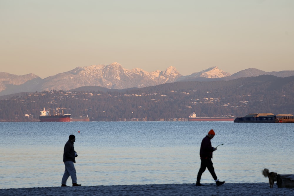 a couple of people walking along a beach with a dog and a mountain in the background